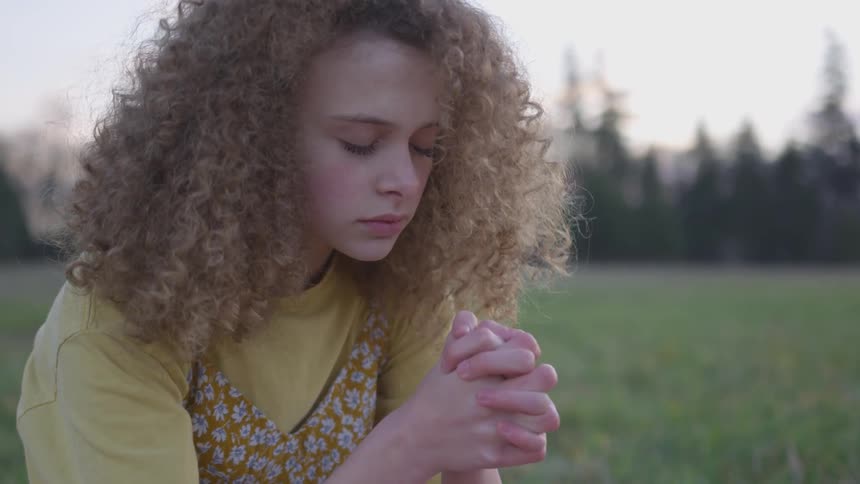 A girl prays while kneeling in a grass field at sunset