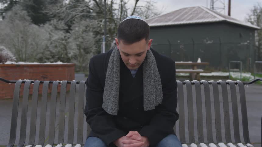Young man in yarmulke sits on park bench with head bowed and hands clasped