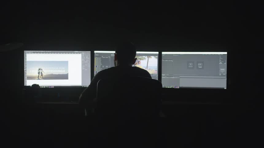 A man looks at three screens in a dark room