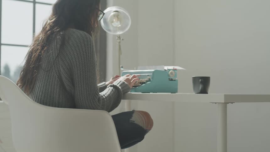 A bespectacled young woman is using an analogue typewriter on a white desk as she sits in a white chair