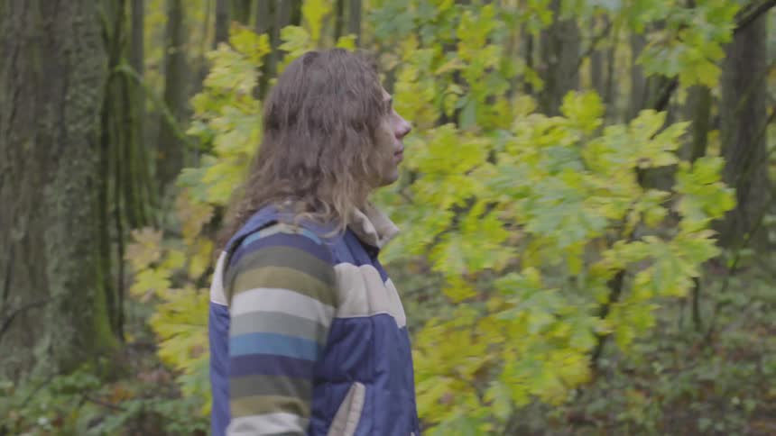 A man with long hair wearing a jacket and a colorful striped shirt walks through the forest on a dirt trail during the midday light