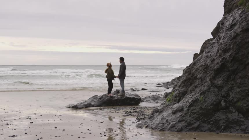 A couple atop a rock holds hands while at the beach
