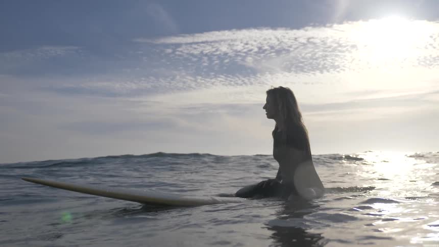 A surfer sits on her surfboard in the ocean and bobs on the waves