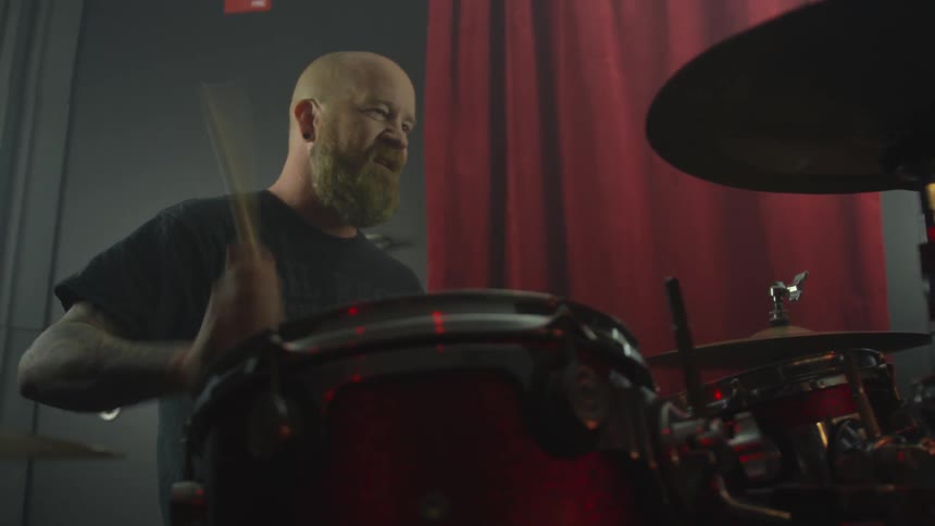 A man plays a drum set in front of a red curtain