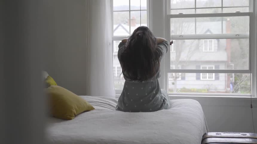 Women fixes her hair in bed in front of a bright window with daylight pouring through on white sheets in a dim room