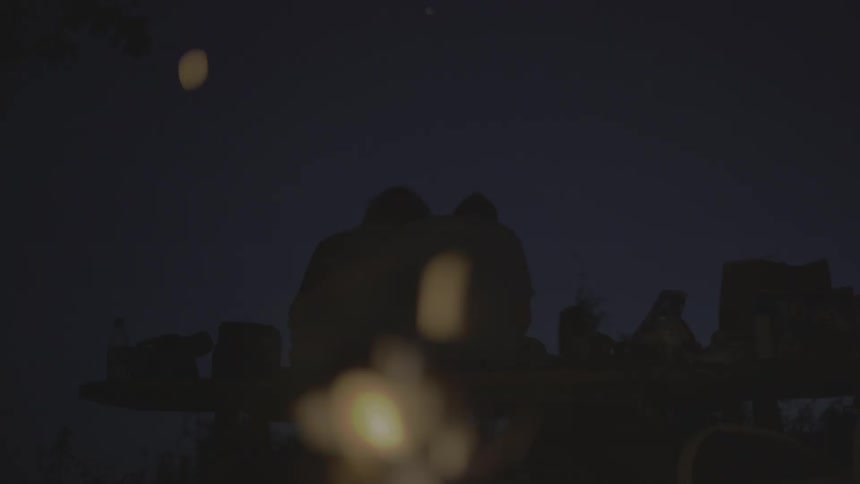 Two people sit embraced at night while a campfire burns behind them