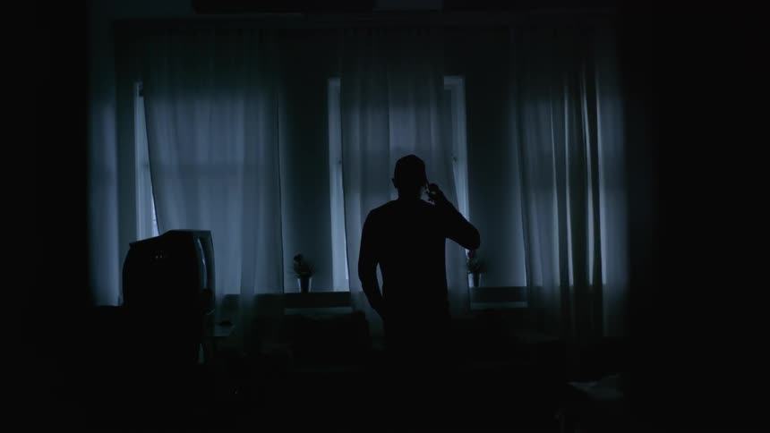 A man walking through a dim front room as he holds a phone up to his ear