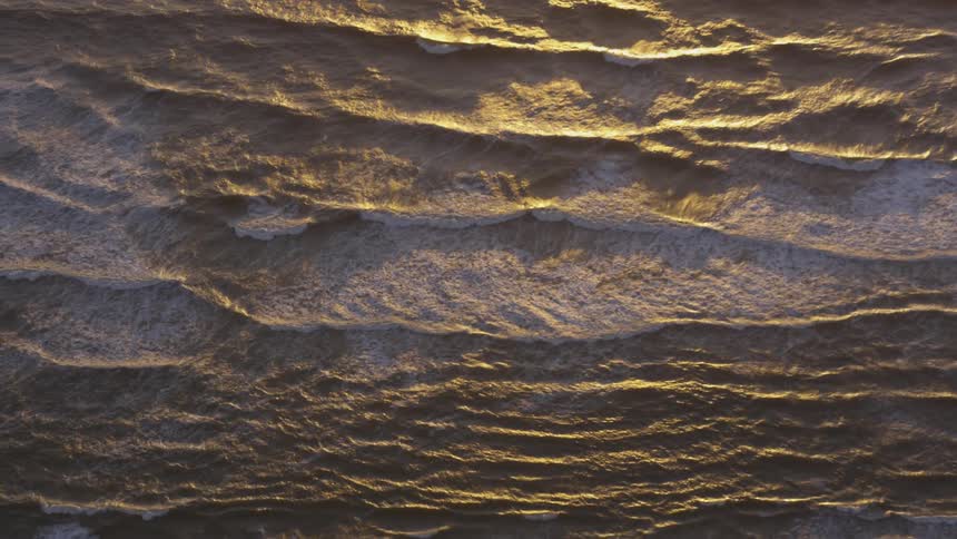A beautiful shot of the waves of an ocean as the sun sets and casts shadows on the waves and white sea foam is seen