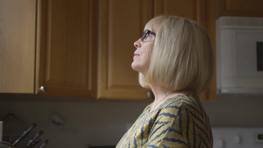 An elderly women in glasses is seen staring out her kitchen window inside her home on a bright sunny day