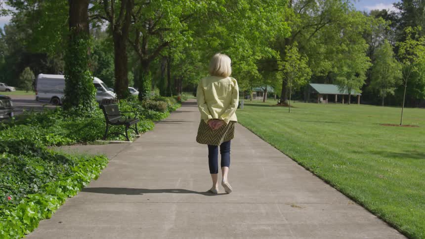 Women walks in slow motion on the sidewalk at the park on a bright sunny day with her hands behind her back and a shadow cast