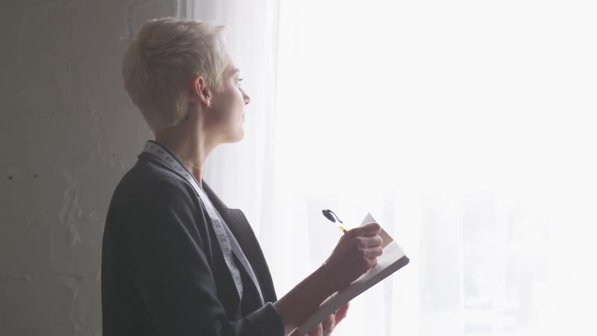 A woman with very short white hair is standing near a window writing on a notepad with a black pen in the sunlight