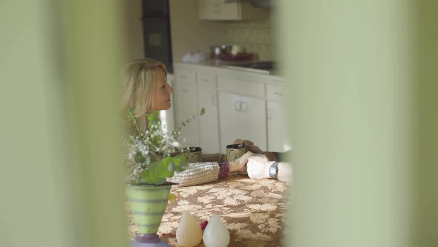 A woman sits in a sunny kitchen across from a man at the table