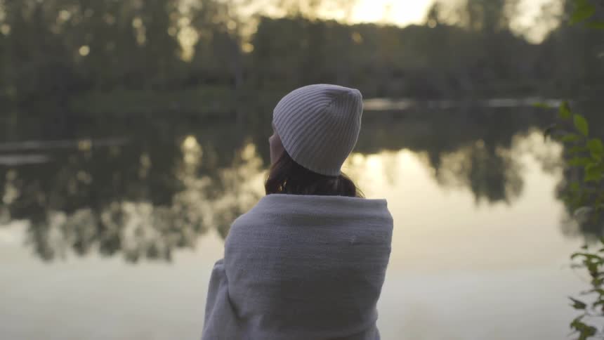 A woman wearing a white hat and wrapped in a white blanket looks out on a river that is reflecting the trees on the other side