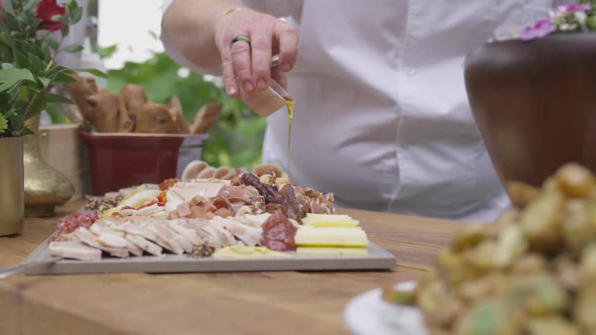 Man pours and drizzles oil on to an arrangement of assorted meats and cheese or butter on a tray on a wooden table outside