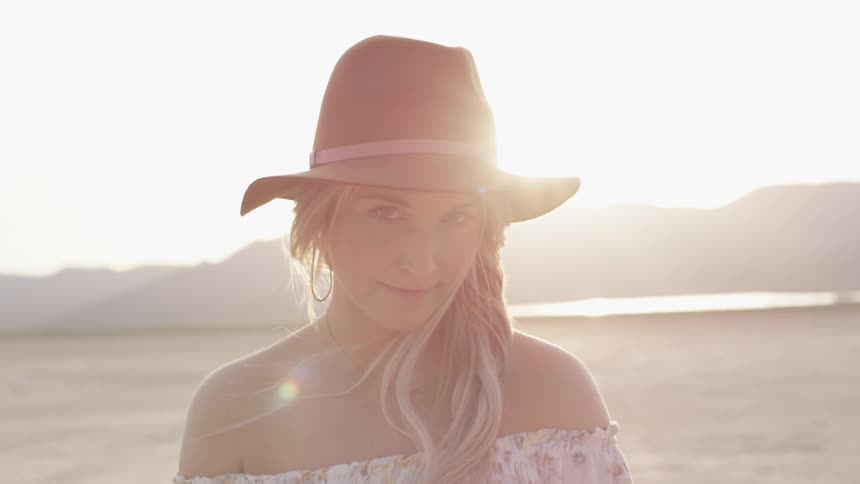 A woman with a hat looks up at the camera while on a beach at dawn