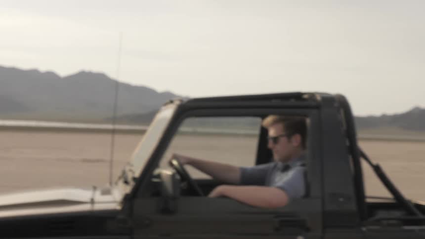 Man leans out door of moving vehicle as it passes another on a wide arid plain