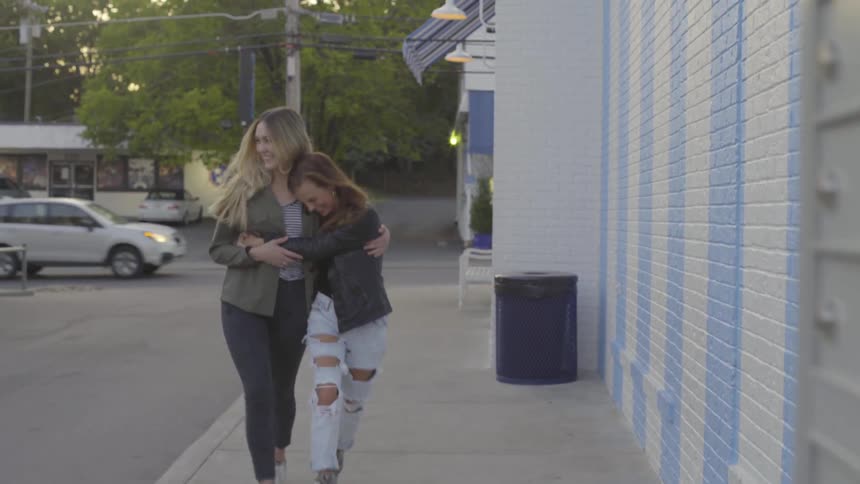Two women smile and hug while walking on a sidewalk at sunset