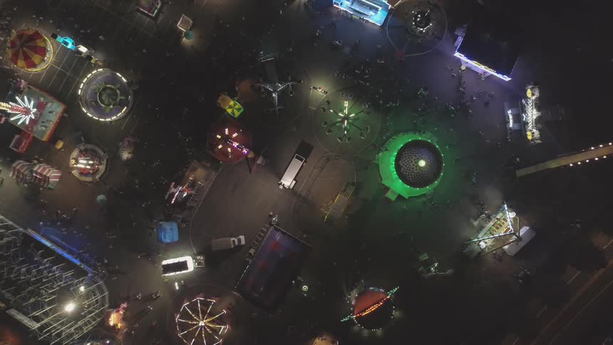 Neon lights adorn the amusement park rides on a dark night with a time lapse of people walking around