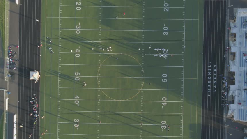 An aerial view of a football field with players walking or entering the grass on a bright sunny day