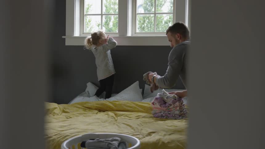 A child stands on the bed as her dad cleans up the mess on the bed on a bright sunny day indoors at home