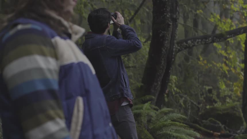 Woman stands nearby as man takes photographs in forest