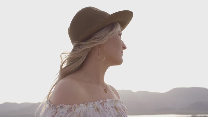 A woman stands out in the open of a desert wearing a white dress and a brown hat on a barren mountain range landscape as the sunset occurs