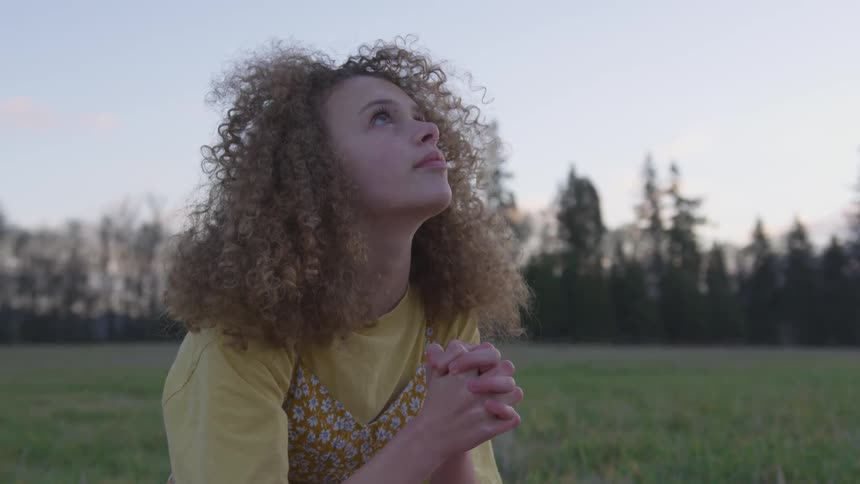 Woman clasps hands and looks up toward sky as she sits in field during the day