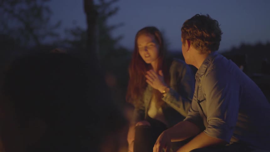 A group of friends sit around a campfire and sing and dance on a dark night