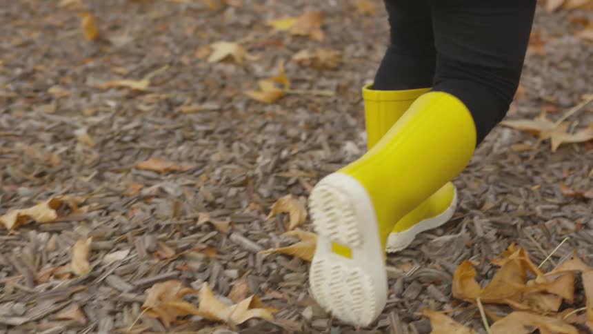A young child walks over bark and autumn leaves while wearing yellow rain boots