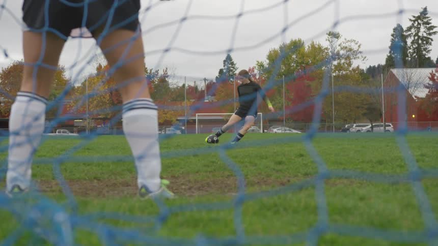 Woman kicking a goal into a soccer net past a goalie