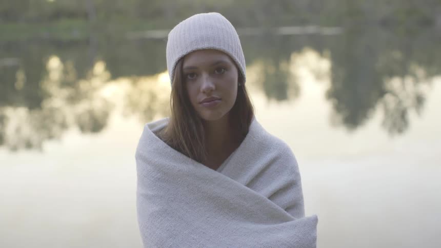 Woman wrapped in towel in front of lake looks up at camera