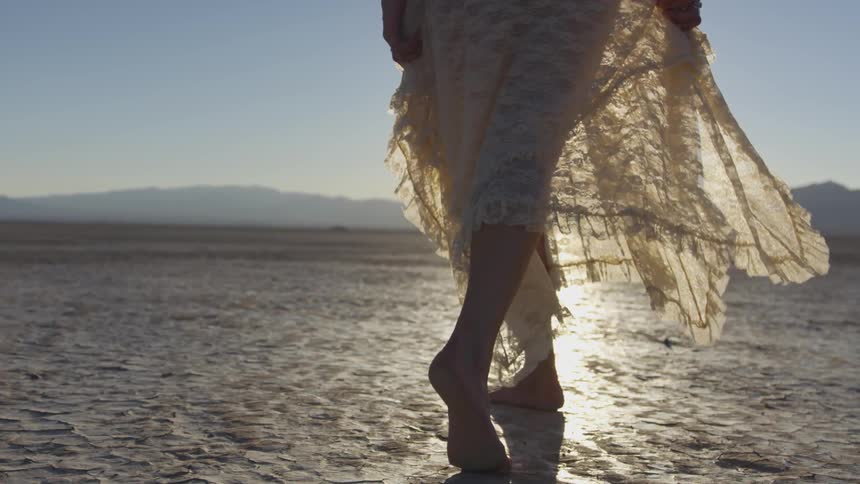 A woman wearing a white dress walking on dry dirt in the sun