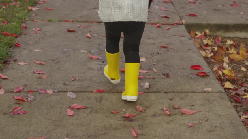 A child in yellow rain boots runs on the sidewalk