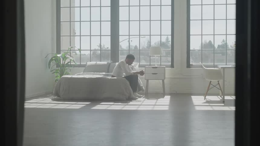 A man sits on a bed in an apartment with large windows