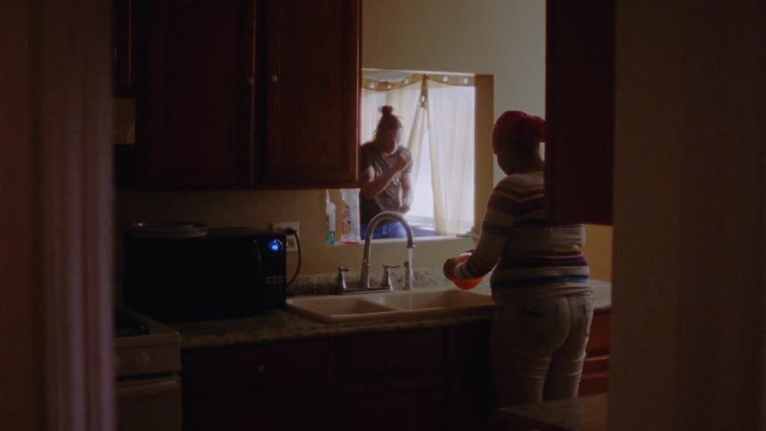 A man is shadow boxing as a woman does dishes