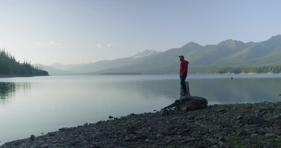 Man wearing hat and red coat stands on rock by lake at sunset