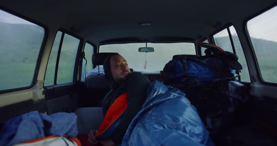 Man in a sleeping back in the back of a vehicle sits up and rubs his eyes