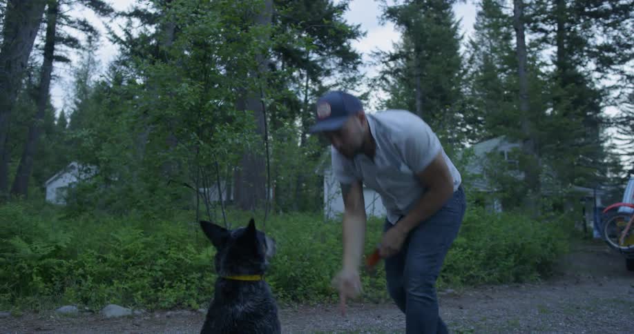 A man in a flat peak cap plays with his dog in a rural area