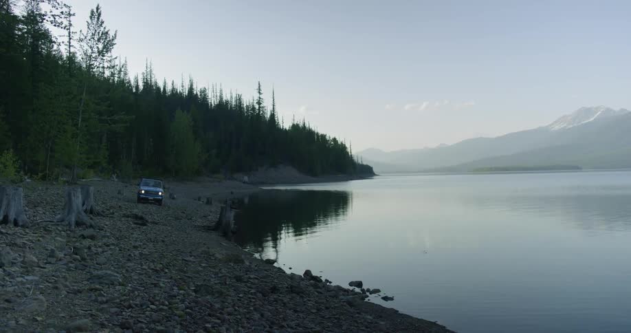 A car drives along the shore of a lake with its headlights on