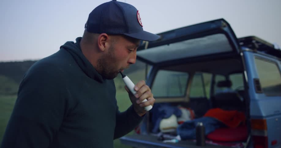 A man brushes his teeth next to a car in a field