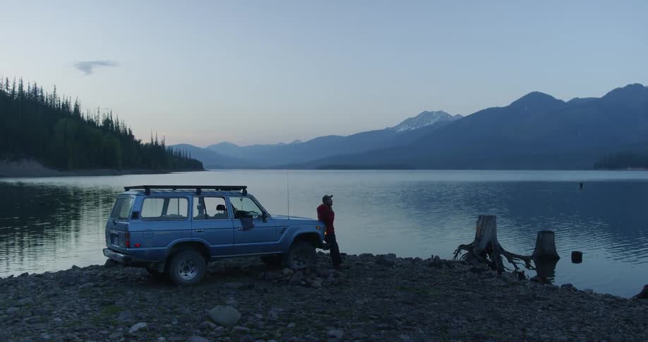 Man leans on hood while looking the water and mountains