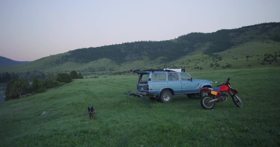 A man unloading a pickup truck on a grassy field with a dog and a motorcycle