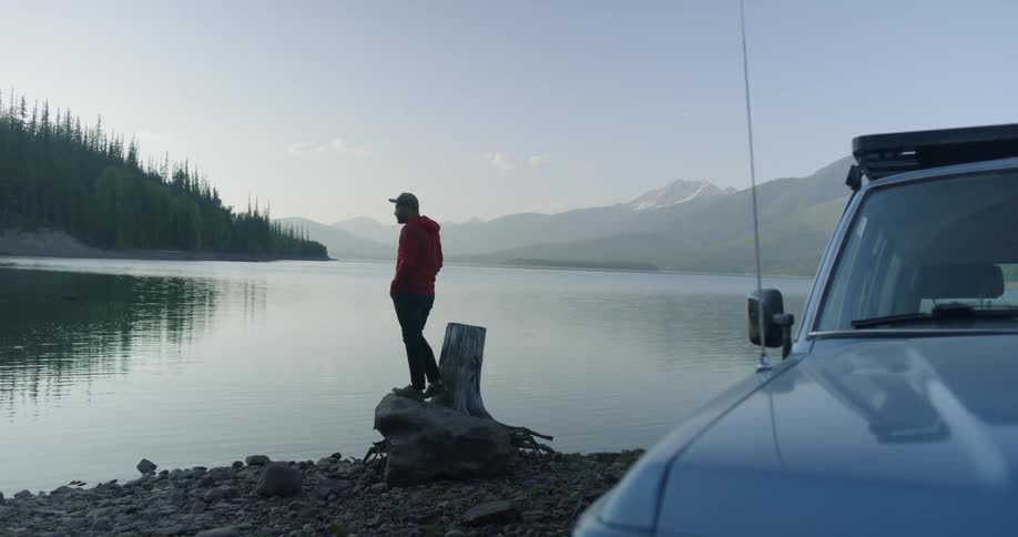 A man stands next to a stump and car on a lake shore