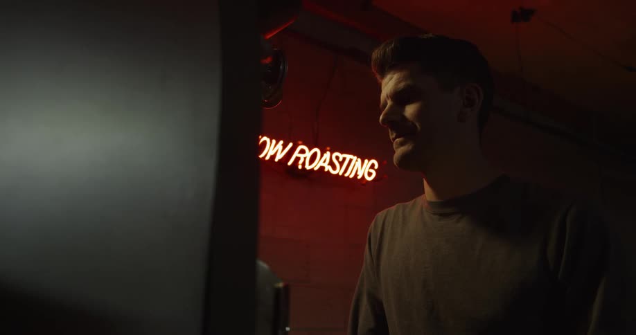 Man stands near large machine in dim light with "now roasting" sign in background
