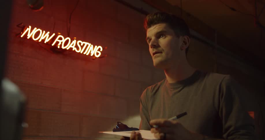 A man takes notes under a neon now roasting sign