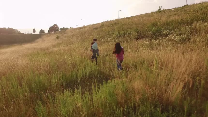 A couple carrying children and walking through tall grass located at the foot of a hill next to a road