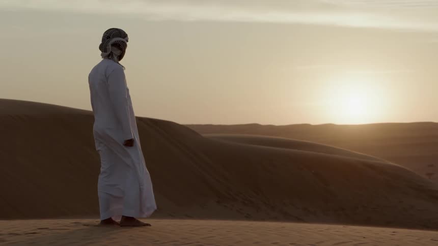 Man in robes stands in desert and looks at sunset