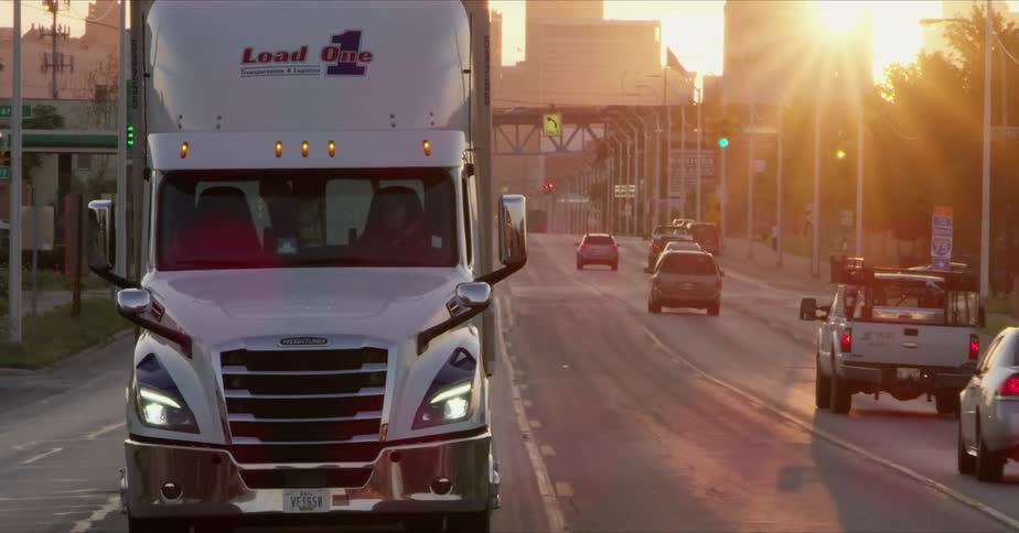 A tractor trailer rides down a six lane highway traveling away from the sun
