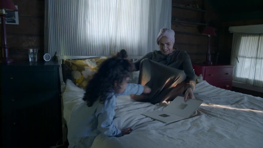 A girl grabs a book from a shelf and brings it to a woman sitting on a bed