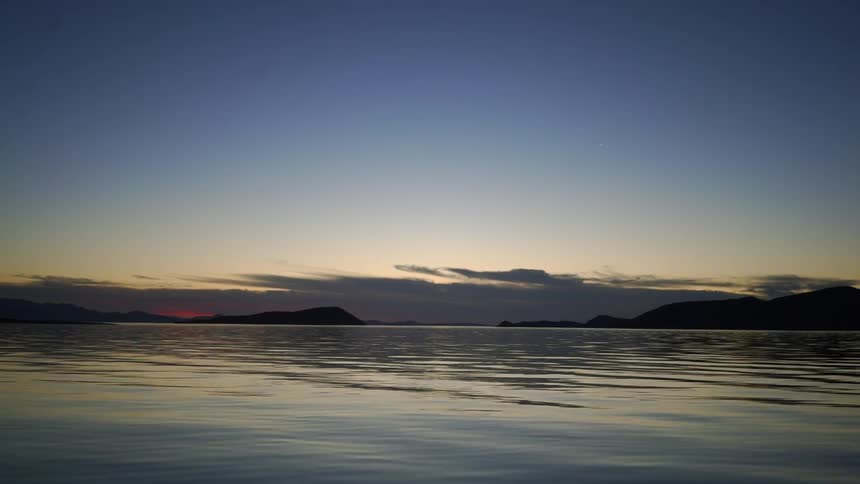 Calm waters are met on the horizon with small hills, orange light and blue skies